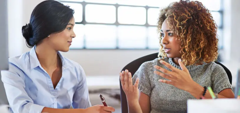 Two women sit a desk in an office and have a conversation 