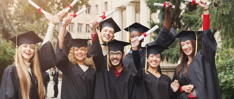 A group of multicultural students holding their diplomas in the air cheering, while wearing a cap and gown with red accents